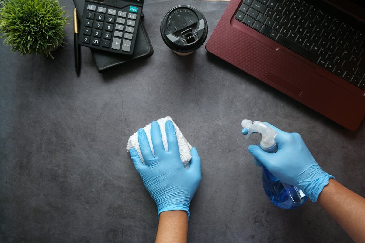 Gloved hands wiping down a desk surface with a cloth and spray bottle